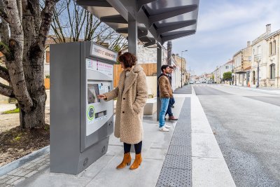 Visuel d'une station de bus express avec au premier plan une femme qui achète son ticket de transport et au second plan deux hommes qui attendent le bus express 